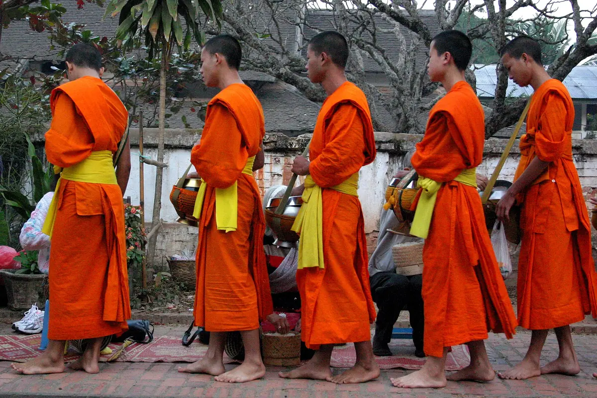 Five monks in orange robes and yellow sashes walk barefoot along a brick pavement with alms bowls, as villagers kneel with offerings.