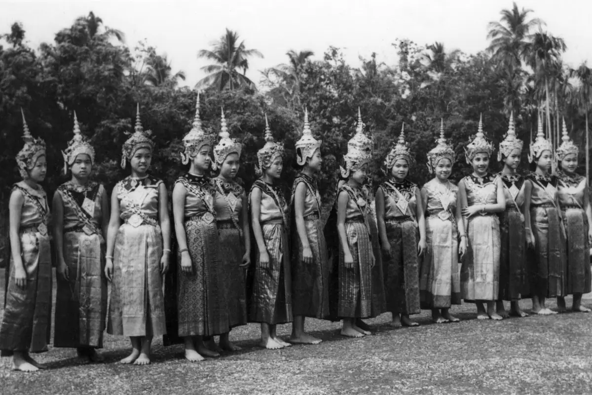 A black-and-white photograph from 1957 of a line of young royal dancers in tall pointed crowns and brocade costumes, standing barefoot on a lawn.