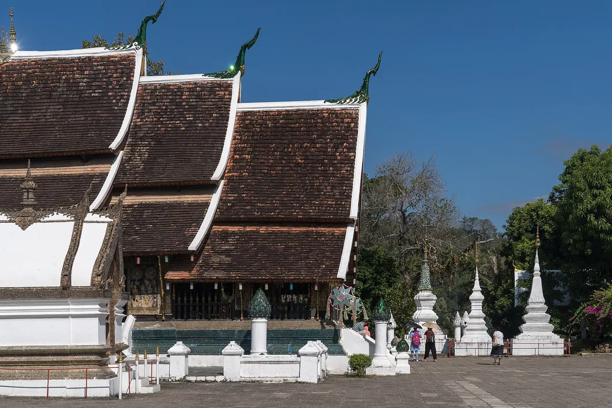 Sweeping tiled roofs of a temple hall at Wat Xieng Thong, with white stupas and visitors in the courtyard under a deep blue sky.